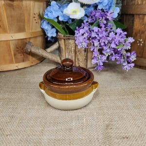 May include: A small, round, two-toned ceramic pot with a lid. The upper portion and lid are a glossy dark brown, while the lower base is a smooth, light cream color. The pot features two small, integrated side handles for easy gripping.