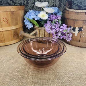 May include: A set of two nested round bowls made of translucent purple glass. The bowls feature smooth, curved sides and a wide rim. The smaller bowl is placed inside the larger bowl, creating a layered appearance with a consistent dark purple hue.