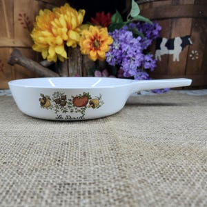 May include: White ceramic skillet with a long handle. The skillet features a decorative border with illustrations of tomatoes, mushrooms, and other vegetables. The words "Le Persil" are written below the vegetables. The skillet is on a burlap surface.