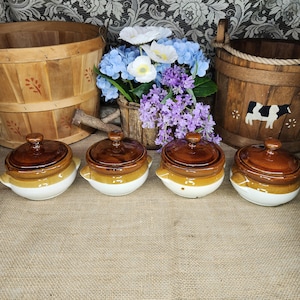 May include: Four small ceramic pots with lids, each featuring a glossy dark brown top and a cream-colored base with two side handles. The pots are arranged in a row in front of two wooden buckets and a bouquet of blue, white, and purple flowers.