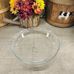 May include: A clear glass pie dish with a circular shape, featuring a fluted rim and two small integrated handles on opposite sides. The dish has a smooth, transparent surface and a shallow basin, designed for baking pies or other culinary creations.