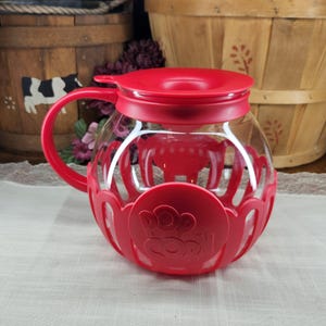 May include: A red popcorn maker with a clear glass bowl and a red handle. The popcorn maker has a red lid and a red base with the word "POP CORN" printed on it. The popcorn maker is sitting on a white surface.