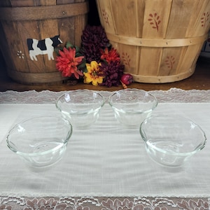 May include: Four clear glass bowls with scalloped edges are arranged on a white lace-trimmed tablecloth. The bowls are in the foreground, with wooden baskets and colorful flowers in the background.