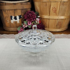 May include: A clear, glass candy dish with a lid. The dish has a textured, diamond-like pattern and a small knob on the lid. The dish is sitting on a white tablecloth. Behind the dish are two wooden baskets and some flowers.