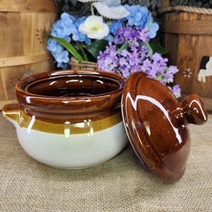 May include: A round ceramic pot with a glossy brown lid and a matching brown rim over a speckled cream-colored base. The pot features a small handle on the side and a knob on the lid, showcasing a two-tone glazed finish with a smooth, reflective surface.