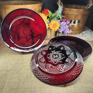 May include: A set of deep red glass plates featuring a pressed geometric pattern with a central starburst design and a diamond-cut border. The plates are circular with a scalloped edge, showcasing a translucent ruby color and intricate relief details.