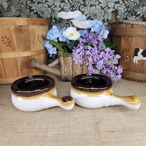 May include: Two ceramic soup crocks with handles, featuring a white base, a brown glazed rim, and a brown accent on the handle tip. The bowls have a smooth, rounded shape and are positioned side by side, showcasing a glossy finish on the interior.