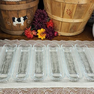 May include: Six clear glass corn-on-the-cob serving dishes with a woven texture. The dishes are rectangular with a curved end and a small handle. The background includes wooden baskets and flowers.