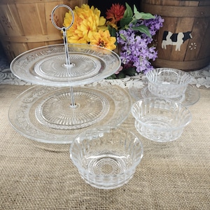 May include: A clear, two-tiered serving tray with a silver handle, and several small clear glass bowls. The tray and bowls have a decorative embossed pattern. The background includes flowers and a wooden basket.