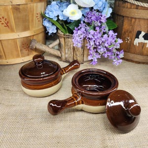 May include: Two ceramic soup crocks with handles and matching lids. Each crock features a two-tone design with a glossy dark brown glaze on the upper portion and lid, transitioning to a smooth cream-colored base on the lower section of the bowl.
