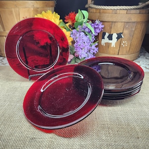 May include: A set of red glass plates with a white rim. The plates are arranged on a burlap surface, with a few plates stacked together. The background includes a wooden basket and colorful flowers.