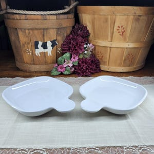 May include: Two white, leaf-shaped serving dishes are displayed on a white lace tablecloth. The dishes have a smooth, glossy finish. In the background are two wooden baskets and a floral arrangement.
