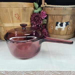 May include: A vintage, ruby-colored glass saucepan with a matching lid. The saucepan has a long handle and is set on a white lace-trimmed cloth. The background includes wooden baskets and purple flowers.