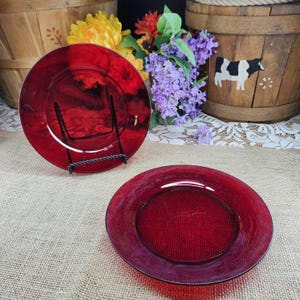 May include: Two ruby red glass plates. One plate is displayed on a black metal stand. The plates have a circular shape with a slightly raised rim. The background includes floral arrangements and a wooden basket.
