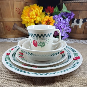 May include: A white ceramic dinnerware set with a green and red farm-themed design. The set includes plates, bowls, and a mug, all featuring the words "Farm Fresh" and "Homemade". The plates and bowls have a green checkered border.
