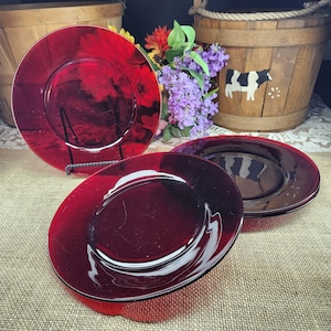 May include: Set of three ruby red glass plates. The plates have a simple, elegant design with a slightly raised rim. The plates are displayed on a burlap surface, with a wooden bucket and flowers in the background.