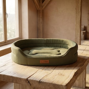 Puede incluir: Una cama para perros verde oliva con un cojín en forma de hueso. La cama está sobre una mesa de madera. La cama tiene un aspecto suave y afelpado. Tiene una pequeña etiqueta rectangular en el frente.