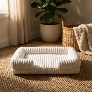 Puede incluir: Una cama rectangular para mascotas de pana blanca con laterales elevados. La cama está sobre una alfombra de yute. Una maceta y una cesta tejida están en el fondo. Las dimensiones de la cama son en centímetros.