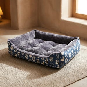 May include: A rectangular pet bed with a plush, gray interior and a patterned exterior. The exterior features a navy blue fabric with a repeating pattern of white cat faces and abstract shapes. The bed sits on a neutral-colored rug.