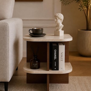 Puede incluir: Una mesa auxiliar de dos niveles con una base de madera oscura en forma de cruz y estantes de color beige claro con aspecto de piedra. El estante superior sostiene una taza de cerámica gris oscuro y un busto blanco. El estante inferior contiene un difusor de vidrio y dos libros etiquetados como CHANEL y VOGUE.