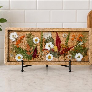 May include: A rectangular wooden tray featuring a collection of pressed dried flowers and foliage embedded in a clear resin surface. The arrangement includes white daisies, green ferns, and reddish-orange leaves, supported by a black metal display stand.