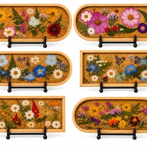 May include: Six rectangular and oval wooden trays featuring pressed dried flowers and greenery embedded in a clear resin base. Each tray is displayed on a black metal stand, showcasing various floral arrangements in shades of yellow, pink, blue, and white.