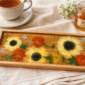 May include: A rectangular wooden tray featuring a clear resin inlay embedded with pressed yellow sunflowers, orange marigolds, and small white flowers. The natural wood grain is visible beneath the floral arrangement, which is framed by a smooth, light-toned wooden border.