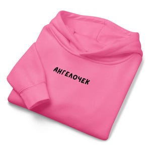 Puede incluir: Una sudadera con capucha rosa brillante doblada con la palabra "АНГЕЛОЧЕК" impresa en negro en la parte delantera. La sudadera con capucha tiene una capucha y mangas largas y está hecha de un material suave.