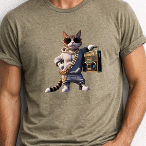 May include: A heathered olive green crew neck t-shirt featuring a graphic of a tabby cat wearing a grey hoodie, dark sunglasses, and a thick gold chain. The cat is holding a vintage-style black boombox with blue speakers in its right paw.