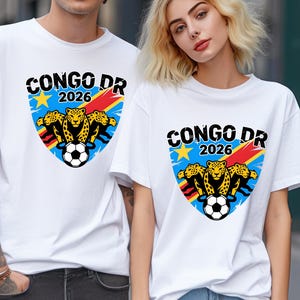 May include: A white t-shirt featuring a graphic of three leopards, a soccer ball, and a shield design with the text CONGO DR 2026. The shield incorporates blue, yellow, and red colors, with a yellow star positioned above the leopards.