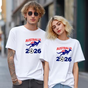 May include: Two people wearing white t-shirts with the word "AUSTRALIA" in red above a blue kangaroo with the Australian flag and a soccer ball, with the number "2026" in blue below. The shirts are short-sleeved.