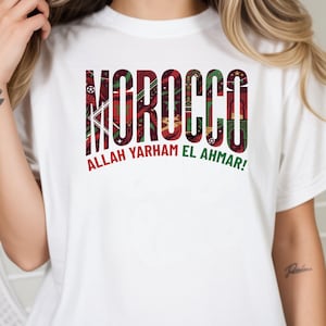 May include: A white short-sleeved t-shirt featuring the word MOROCCO in large block letters filled with a red, green, and black patterned design. Below the text, the phrase ALLAH YARHAM EL AHMAR is printed in red and green capital letters.
