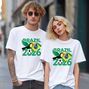 May include: White t-shirts with a Brazil 2026 graphic. The design features a toucan, a soccer ball, and the Brazilian flag colors. The word "BRAZIL" is in green, and the year "2026" is below the toucan.