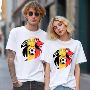 May include: White t-shirts featuring a graphic of a lion's head with the word "BELGIUM" and the year "2026". The lion design incorporates the colors of the Belgian flag and a soccer ball. The shirts are short-sleeved.