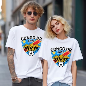 May include: White t-shirts featuring a shield design with the text "CONGO DR 2026". The design includes a soccer ball, three leopards, and a star. The shirts are worn by two people.