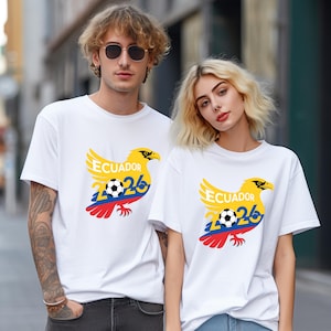 May include: White t-shirts with a yellow eagle design, the word "Ecuador", and the number "26" in blue. A soccer ball is in the center of the design. The eagle has red, yellow, and blue accents. The shirts are worn by two people.