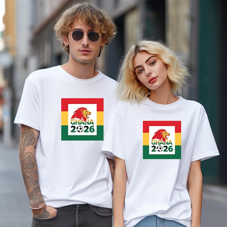 May include: Two people wearing white t-shirts. Each shirt features a square design with the Ghana flag colours: red, yellow, and green. The design includes a lion, the word "Ghana", the year "2026", and a football.