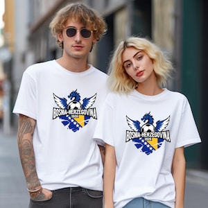 May include: White t-shirts featuring a graphic design with a blue dragon, soccer ball, and the text "BOSNIA-HERZEGOVINA 2026". The design incorporates the colors of the Bosnian flag. The shirts are worn by two people.