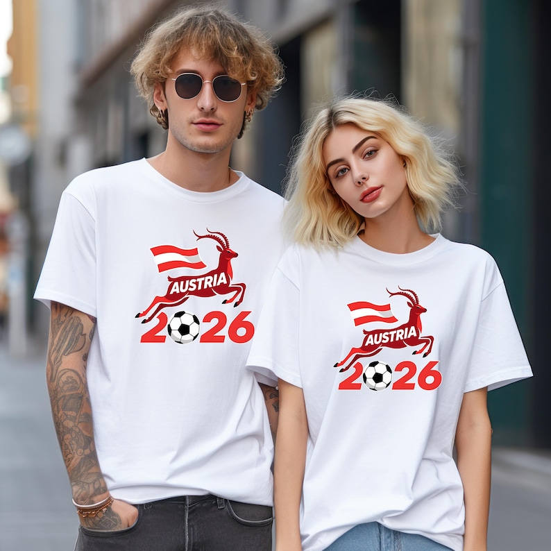 May include: White t-shirts with a red and black graphic featuring a deer, the Austrian flag, a soccer ball, and the text "AUSTRIA 2026". The shirts are worn by two people.
