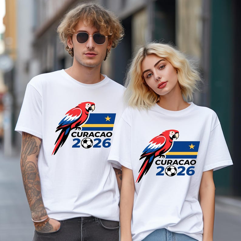 May include: White t-shirts with a parrot design in red, blue, and black, a football, and the text "CURACAO 2026". The shirts are worn by two people.