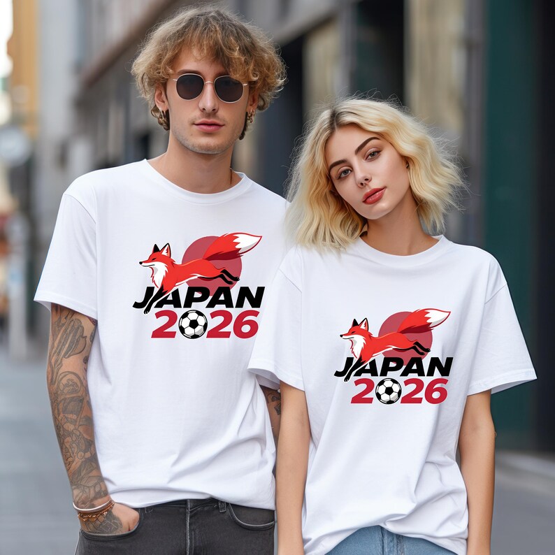 May include: White t-shirts with a graphic of a red fox leaping in front of a red circle, with the words "JAPAN 2026" in black and red. The design includes a football. The shirts are short-sleeved.