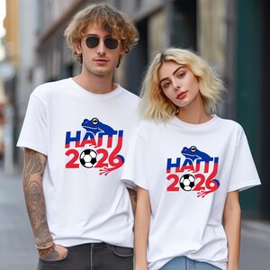 May include: White t-shirts with a graphic design featuring a blue frog, the word "HAITI", the number "2026", and a soccer ball. The design incorporates red and blue colours. The shirts are worn by two people.
