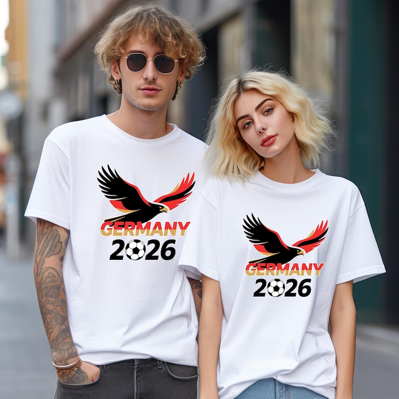 May include: White t-shirts with a black eagle design, red and yellow accents, the word "GERMANY", the year "2026", and a soccer ball graphic. The shirts are worn by two people.