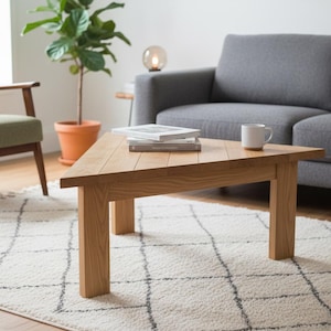 Peut inclure: Une table basse triangulaire en bois avec une finition brun clair et un grain de bois visible. La table présente trois pieds robustes et une surface plane. Une pile de trois livres et une tasse en céramique blanche sont posées au centre.
