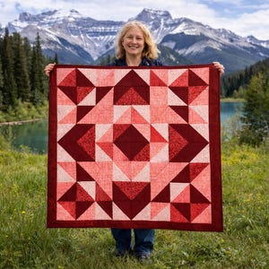 Peut inclure: Une courtepointe faite à la main avec un motif géométrique dans les tons de rouge et de rose. La courtepointe présente des carrés, des triangles et des losanges, avec une bordure rouge foncé. La courtepointe est tenue devant un arrière-plan de montagne et de lac.