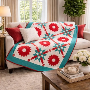 Peut inclure: Une courtepointe crème avec un motif d'éclat d'étoile rouge et turquoise est drapée sur un canapé beige. La courtepointe a une bordure turquoise et est entourée de coussins rouges. Un coussin blanc et une table d'appoint en bois avec une lampe sont également visibles.