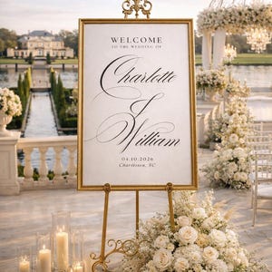 Pode incluir: Uma placa de boas-vindas dourada para um casamento, com os nomes Charlotte e William, e a data 04.10.2026. A placa está em um cavalete dourado, rodeada por flores brancas e velas. O fundo mostra um local ao ar livre cênico.
