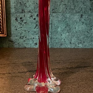 May include: A tall, slender, ruby-red glass vase with a clear, flared base. The vase has a twisted design and a delicate, petal-like base, creating an elegant and artistic appearance. The vase is likely for decorative purposes.