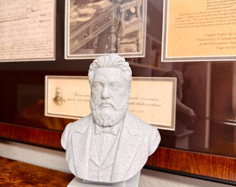 May include: A light gray bust of a man with a beard and bow tie, mounted on a square pedestal. The bust is in front of framed historical documents and a portrait. The bust is made of a speckled material.