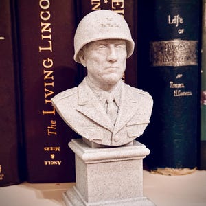 May include: A light gray bust of a military figure wearing a helmet and uniform, mounted on a square pedestal. The bust is set against a backdrop of dark brown and green books.